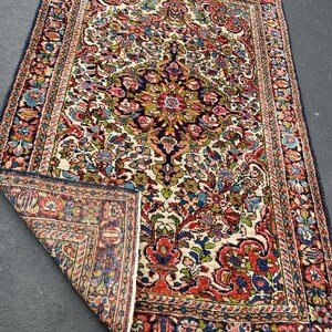 May include: A rectangular, vintage rug with a floral pattern in shades of red, blue, green, and cream. The rug has a dark blue border and a detailed central design. The corner is folded back, revealing the rug's construction.