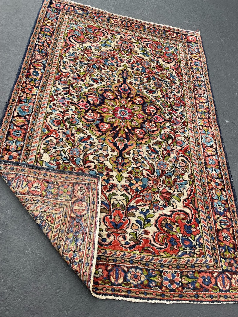 May include: A rectangular, vintage rug with a floral pattern in shades of red, blue, green, and cream. The rug has a dark blue border and a detailed central design. The corner is folded back, revealing the rug's construction.