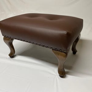 May include: A brown leather ottoman with a rectangular top and two button tufts. The ottoman has four curved wooden legs and is trimmed with silver-colored nailhead studs.