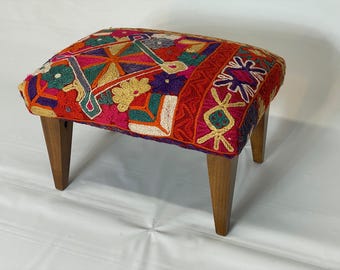 May include: A small, rectangular footstool with a vibrant, embroidered top. The cushion features a patchwork design in red, pink, yellow, green, and purple. The wooden legs are tapered and angled, providing a sturdy base for this decorative piece.