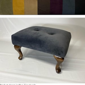 Pode incluir: Um pufe estofado em camurça azul-acinzentado com duas tufos de botões. O pufe tem quatro pernas de madeira escura curvas. A parte superior da imagem mostra uma amostra de cores de diferentes cores de tecido.