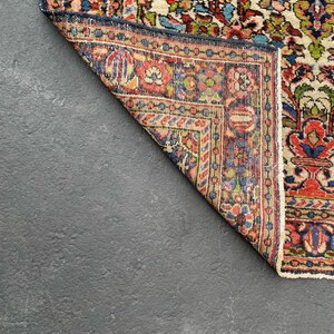 May include: A section of a vintage rug with a detailed floral pattern. The rug features a cream-colored field with red, blue, and green floral motifs. The border is a deep blue with intricate designs. The rug is partially folded, revealing the underside.