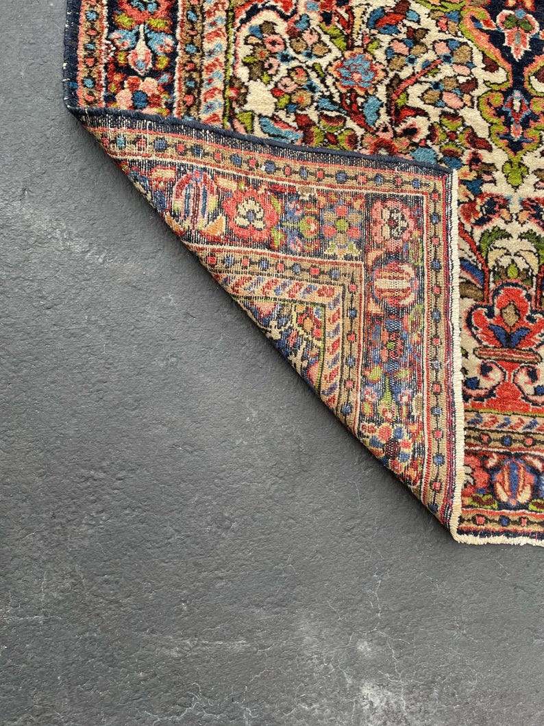 May include: A section of a vintage rug with a detailed floral pattern. The rug features a cream-colored field with red, blue, and green floral motifs. The border is a deep blue with intricate designs. The rug is partially folded, revealing the underside.