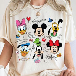 Puede incluir: Camiseta color crema con un gráfico colorido de personajes de Disney, incluyendo Mickey Mouse, Minnie Mouse, Donald Duck, Daisy Duck, Goofy y Pluto. La camiseta tiene el texto "Mickey Mouse" y firmas de personajes.