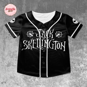 A black baseball jersey with white text and graphics, including a jack Skellington logo, is displayed against a concrete background.
