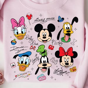 A pink sweatshirt with various Disney characters and logos printed on it, including Mickey Mouse, Donald Duck, Goofy, Minnie Mouse, and Pluto.