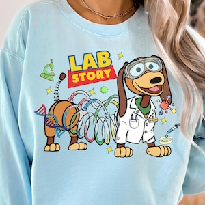 Op de afbeelding: Lichtblauw sweatshirt met een cartoon teckel met een laboratoriumbril en een labjas. De teckel is verbonden met een reeks kleurrijke buizen en wetenschappelijke illustraties. De tekst "LAB STORY" wordt weergegeven in een speels lettertype.