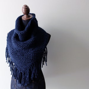 May include: A navy blue knitted cowl with fringe. The cowl is displayed on a wooden mannequin.