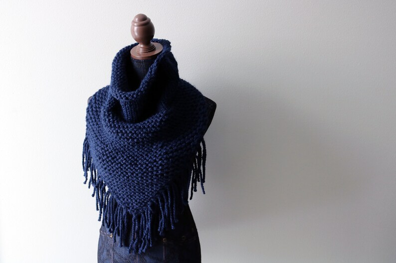 May include: A navy blue knitted cowl with fringe. The cowl is displayed on a wooden mannequin.