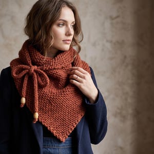 May include: A chunky knit triangular scarf in a burnt orange color. The scarf features a side tie closure with a bow detail and two light-colored wooden beads at the ends of the ties. The fabric has a visible garter stitch texture throughout.