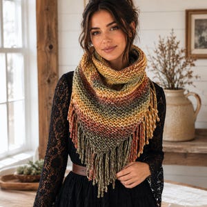 May include: A triangular knit scarf featuring a variegated color palette of mustard yellow, burnt orange, olive green, and cream. The scarf has a textured garter stitch pattern and is finished with long, thick yarn fringe along the bottom edge.