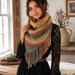 May include: A triangular knit scarf featuring a variegated color palette of mustard yellow, burnt orange, olive green, and cream. The scarf has a textured garter stitch pattern and is finished with long, thick fringe along the bottom edges.