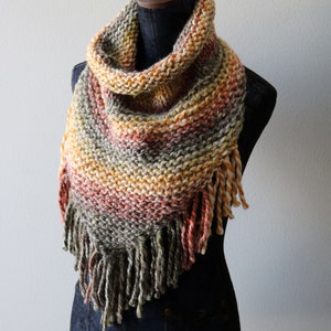 May include: A hand-knitted triangular cowl scarf featuring a variegated yarn pattern in shades of olive green, burnt orange, yellow, and cream. The scarf has a textured garter stitch construction and is finished with long, thick twisted fringe.