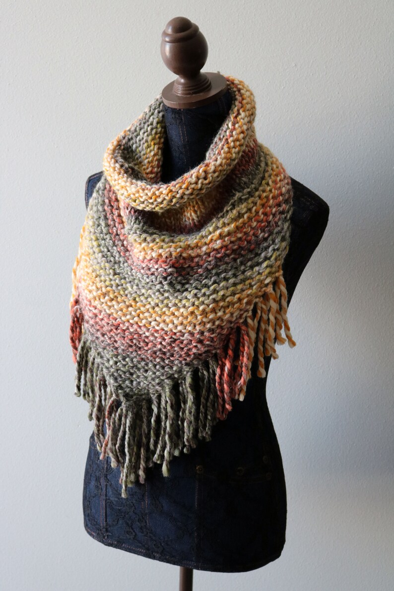 May include: A hand-knitted triangular cowl scarf featuring a variegated yarn pattern in shades of olive green, burnt orange, yellow, and cream. The scarf has a textured garter stitch construction and is finished with long, thick twisted fringe.