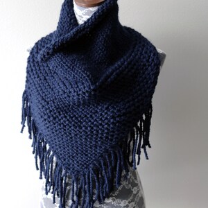 May include: A navy blue knitted cowl neck scarf with fringe. The scarf is displayed on a white lace-covered mannequin.
