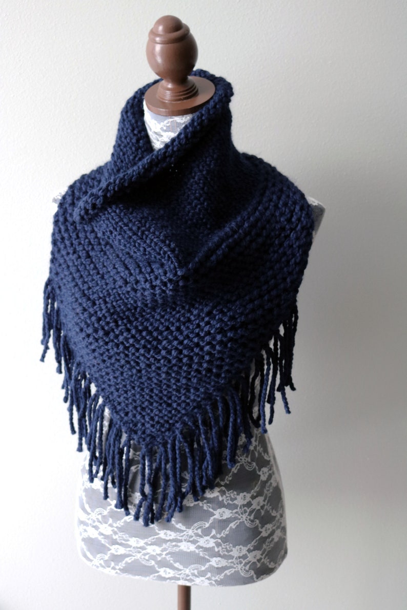 May include: A navy blue knitted cowl neck scarf with fringe. The scarf is displayed on a white lace-covered mannequin.
