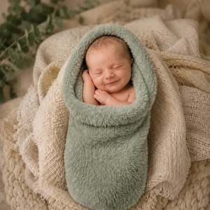 May include: A newborn baby with a gentle smile is wrapped in a soft, sage green fuzzy swaddle. The swaddle features a hood that frames the baby's face, and the infant is resting with hands tucked near the chin, creating a cozy and peaceful appearance.