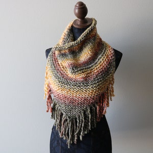 May include: A triangular knit scarf featuring a variegated color palette of olive green, burnt orange, mustard yellow, and cream. The scarf is constructed with a chunky garter stitch pattern and is finished with long, thick fringe along the bottom edge.