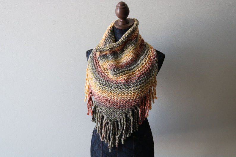 May include: A triangular knit scarf featuring a variegated color palette of olive green, burnt orange, mustard yellow, and cream. The scarf is constructed with a chunky garter stitch pattern and is finished with long, thick fringe along the bottom edge.