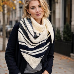 Könnte beinhalten: Ein dreieckiger Strickschal mit einem Color-Blocking-Muster aus cremefarbenen, dunkelgrauen und marineblauen horizontalen Streifen. Der Schal hat eine strukturierte kraus rechts gestrickte Oberfläche und ist um den Hals drapiert.