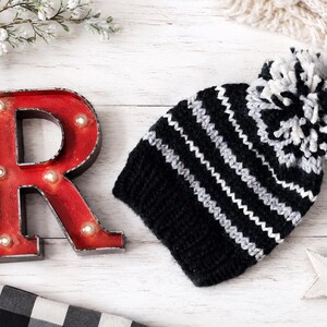 May include: A knit beanie featuring horizontal stripes in black, white, and gray with a matching multicolored pom-pom on top. The hat has a ribbed cuff at the base and is constructed from thick yarn, showcasing a textured pattern throughout the design.