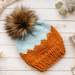 May include: A knit beanie featuring a two-tone colour block design with a light blue upper section and a burnt orange lower section. The hat is topped with a large, fluffy brown faux fur pom-pom and has a ribbed texture along the bottom edge.