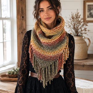 May include: A triangular knit scarf featuring a variegated color palette of mustard yellow, burnt orange, olive green, and cream. The scarf has a textured garter stitch pattern and is finished with long, thick yarn fringe along the bottom edge.