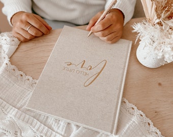 May include: A beige fabric-covered hardcover journal with the gold foil text Hello Little Love printed on the front cover. The journal is positioned on a wooden surface next to a white lace-trimmed cloth and a decorative vase of dried flowers.