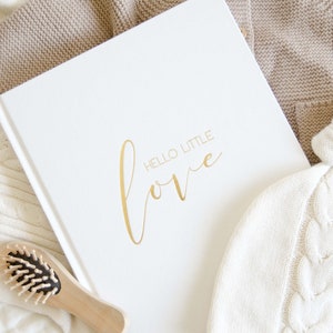 May include: A white book with the words 'HELLO LITTLE love' in gold lettering. The book is surrounded by white and beige knitted baby clothes and a wooden hairbrush.