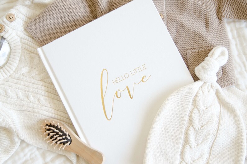 May include: A white book with the words 'HELLO LITTLE love' in gold lettering. The book is surrounded by white and beige knitted baby clothes and a wooden hairbrush.