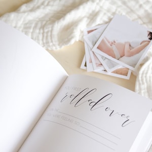 May include: A white baby memory book with a black inscription that reads 'You first rolled over' with lines below for writing the date and how the baby was feeling. The book is open to a blank page. A stack of white photos is on the right side of the book.
