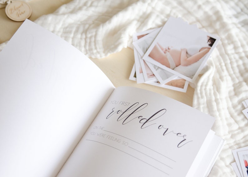 May include: A white baby memory book with a black inscription that reads 'You first rolled over' with lines below for writing the date and how the baby was feeling. The book is open to a blank page. A stack of white photos is on the right side of the book.