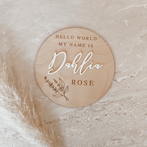 May include: A round wooden sign with the text "Hello World My Name Is Dahlia Rose" in white lettering. The sign has a floral design in brown.