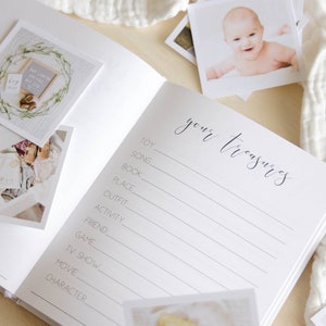 May include: A white baby memory book with the words 'your treasures' written in black cursive script. The book has a list of categories for recording memories such as toy, song, book, place, outfit, activity, friend, game, TV show, movie, and character. There are blank lines next to each category for writing in memories. The book has several photos of a baby in it.