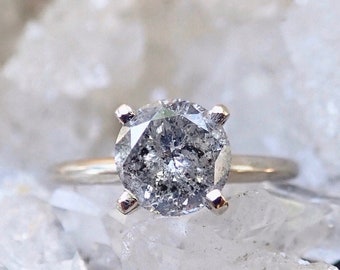 Puede incluir: Un anillo de compromiso solitario con un gran diamante redondo gris con aspecto moteado. El diamante está engastado en una banda de oro con cuatro garras. El anillo se muestra sobre un fondo blanco borroso.