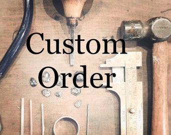 May include: A close-up of a workbench with tools and materials for making custom jewelry. The tools include a hammer, a chisel, a ruler, and a pair of pliers. There are also several small diamonds and a ring band on the workbench. The text "Custom Order" is written in large, bold letters across the image.