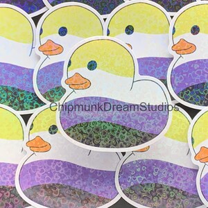 May include: A set of iridescent stickers featuring a cartoon rubber duck with a yellow head, white body, and orange beak. The duck is decorated with a purple, white, and yellow stripe pattern, representing the non-binary pride flag. The background of the stickers is a pattern of iridescent hearts.