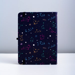 May include: A dark blue notebook cover with a constellation pattern. The constellations are outlined in pink, blue, green, and yellow. The constellations are labeled with their astrological signs.