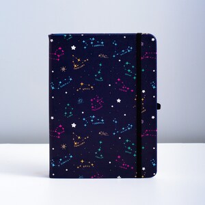 May include: A dark blue notebook with a constellation pattern. The constellations are outlined in pink, yellow, and blue, and the names of the constellations are written in white. The notebook has a black elastic band closure.