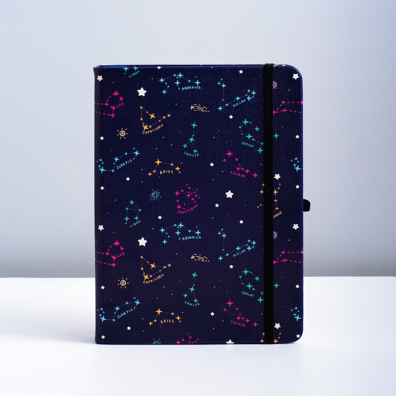 May include: A dark blue notebook with a constellation pattern. The constellations are outlined in pink, yellow, and blue, and the names of the constellations are written in white. The notebook has a black elastic band closure.
