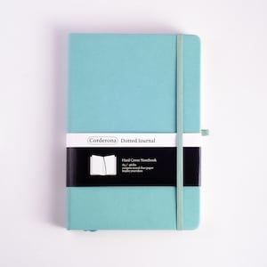 Puede incluir: Un cuaderno de tapa dura punteado de color azul claro con una etiqueta en blanco y negro que dice "Corderona Dotted Journal" y "Hard Cover Notebook As/g6ahs 100gsm wood-free paper Inspire your ideas".