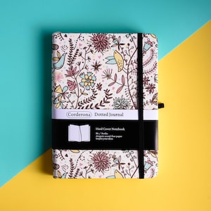 Puede incluir: Un cuaderno de tapa dura con un diseño floral y un cierre elástico negro. La cubierta tiene un fondo crema con un patrón de flores, hojas y pájaros en tonos rosa, amarillo y azul. El texto "Corderona Dotted Journal" está impreso en la cubierta.