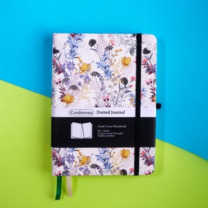 Könnte beinhalten: Ein Notizbuch mit Hartcover und floralem Muster in Gelb, Lila und Blau auf weißem Hintergrund. Es hat einen schwarzen Gummibandverschluss und ein Lesezeichen. Der Text "Corderona Dotted Journal" und "Hard Cover Notebook" sind sichtbar.