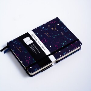 May include: A navy blue hard cover dotted journal with a constellation pattern. The cover features a black and white label with the text "Corderona Dotted Journal" and "Hard Cover Notebook".