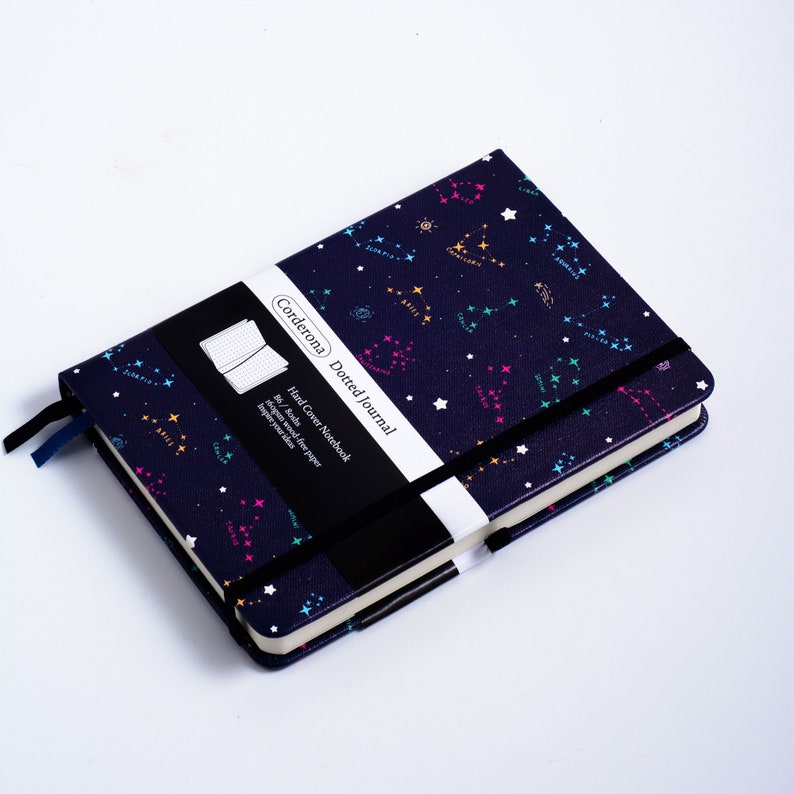 May include: A navy blue hard cover dotted journal with a constellation pattern. The cover features a black and white label with the text "Corderona Dotted Journal" and "Hard Cover Notebook".