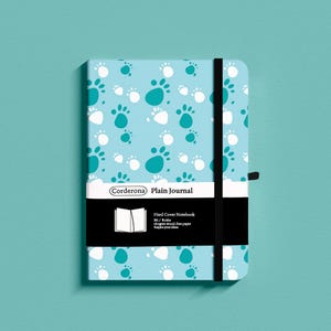 May include: A light blue hard cover notebook with a paw print pattern in white and teal. The notebook has a black elastic band closure and a black band with the text "Corderona Plain Journal". The notebook is 14 cm wide and 21 cm tall.