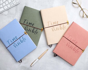 Op de afbeelding: Vier notitieboekjes in pastelkleuren: blauw, groen, beige en roze. Elk notitieboekje heeft de woorden "Time Visible" in reliëf op de kaft en is vastgezet met een elastiek. Een pen en een bril zijn ook zichtbaar.