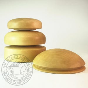 May include: Four light-coloured wooden hat blocks. Three are stacked, and the fourth is beside them. The blocks are different sizes and shapes, with rounded tops and flat bases. The logo "HAT BLOCKS AUSTRALIA MAKING BLOCKS BY DESIGN" is visible.