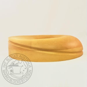 Pode incluir: Um bloco de chapéu de madeira de cor clara com uma forma ovalada e curva. O bloco de chapéu tem um acabamento liso e polido e é projetado para chapelaria. A base do bloco de chapéu tem um logotipo circular com o texto "HAT BLOCKS AUSTRALIA MAKING BLOCKS BY DESIGN."