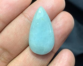 May include: A teardrop-shaped, aquamarine gemstone with a polished surface. The gem has a pale blue color and a smooth, rounded shape. The gemstone is held in a hand, with the background out of focus.